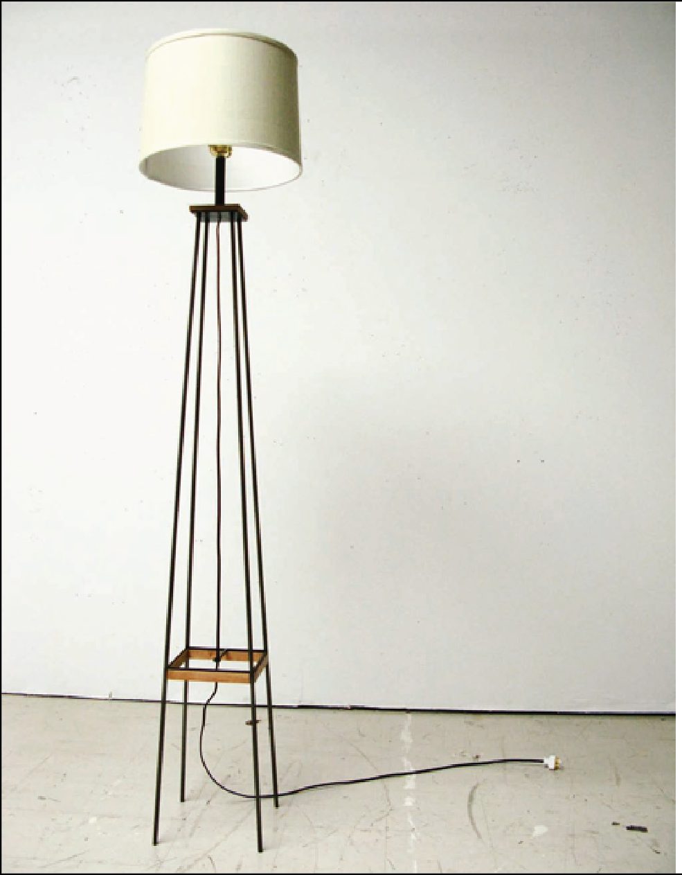 A BILT floor lamp with a slender blackened steel frame, wooden collar and pale drum shade, photographed against a bare studio wall.